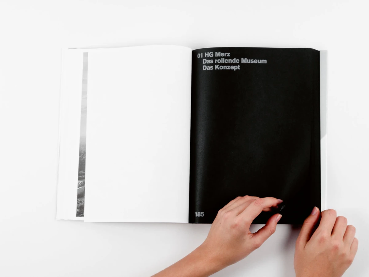 Hands turning a page in an open book; right page is black with white text, left page white on a white surface.