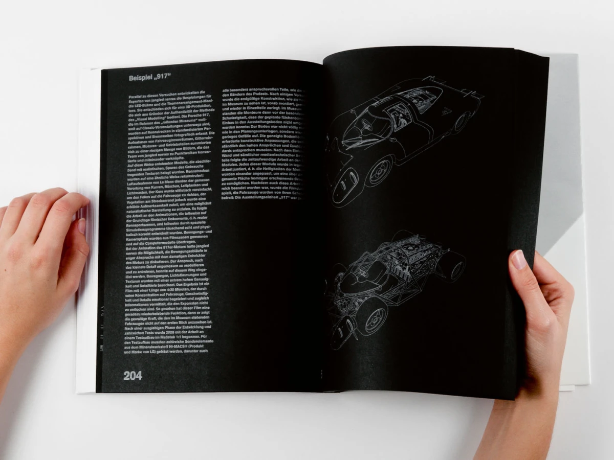Two hands hold an open magazine displaying white car blueprints on black pages against a white surface.