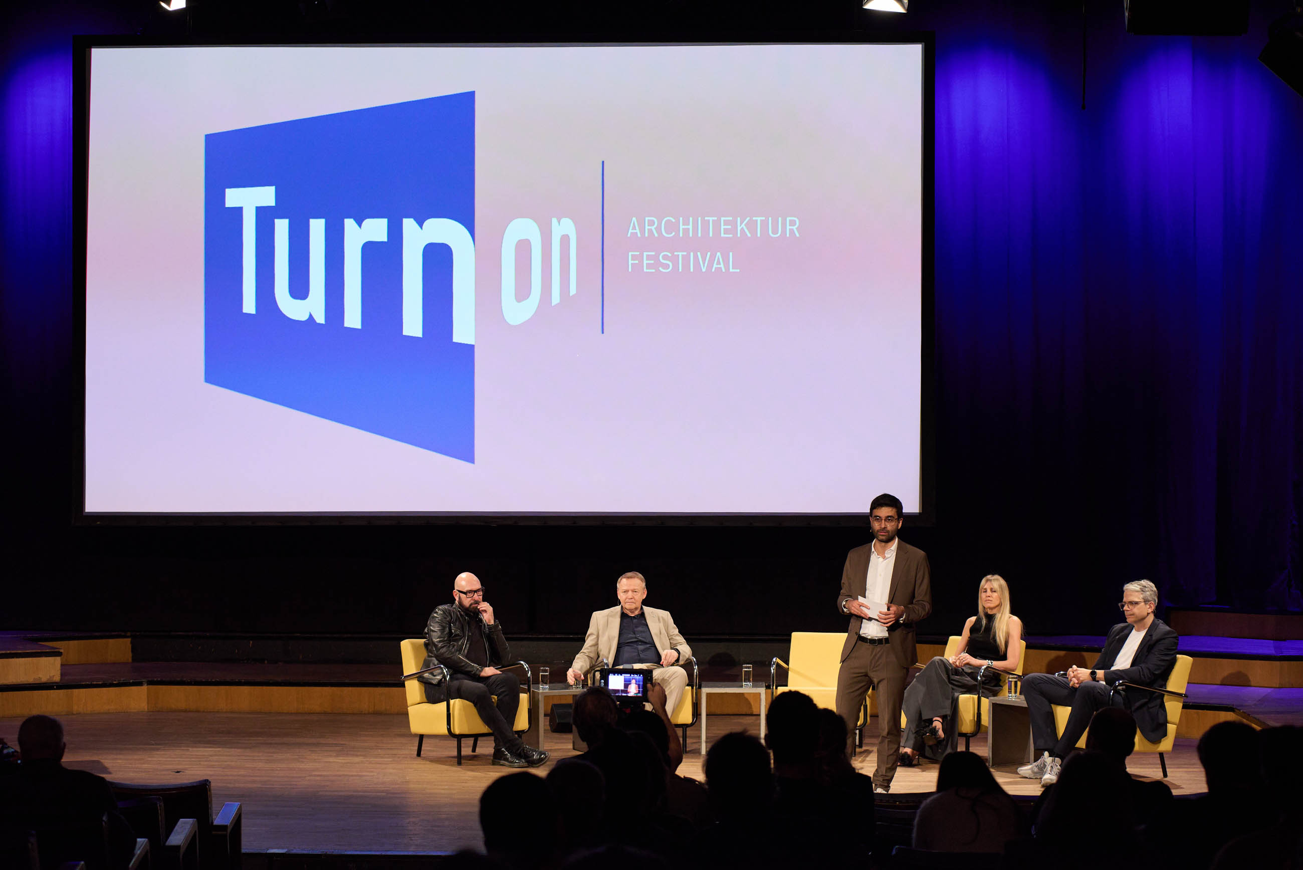 Architecture festival stage panel: five panelists seated, one man standing and speaking; large screen displays Turn On logo.