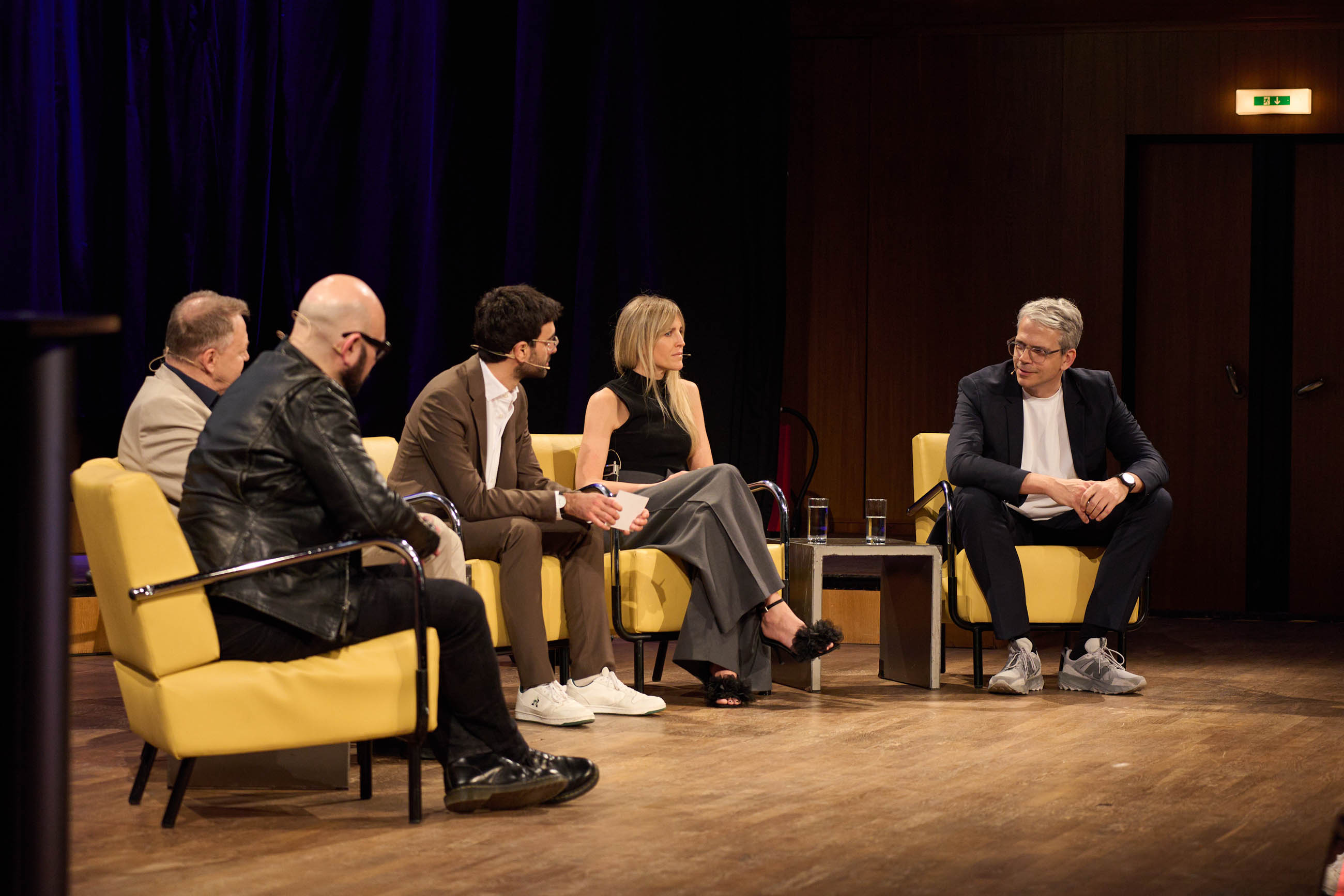 Five panelists seated in yellow chairs on a stage, engaged in discussion; small table with water glasses between them, dark curtain backdrop.