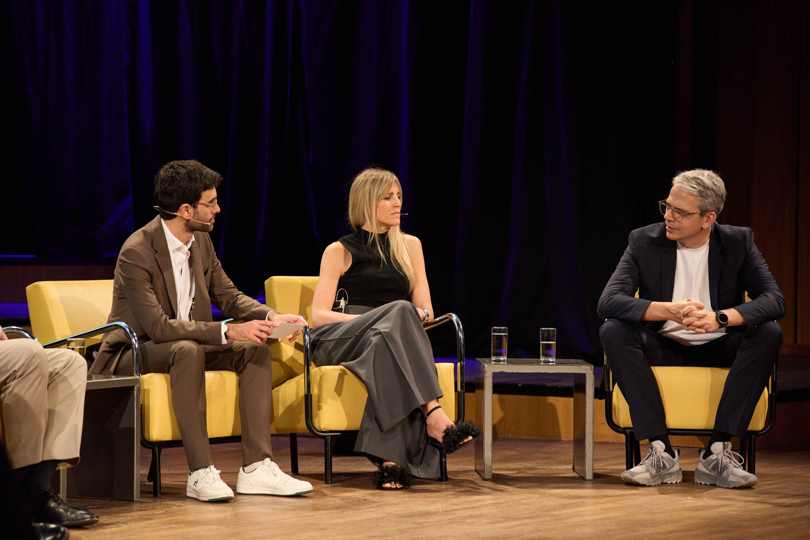 Three panelists seated on yellow chairs on a stage with dark blue curtains, conversing.
