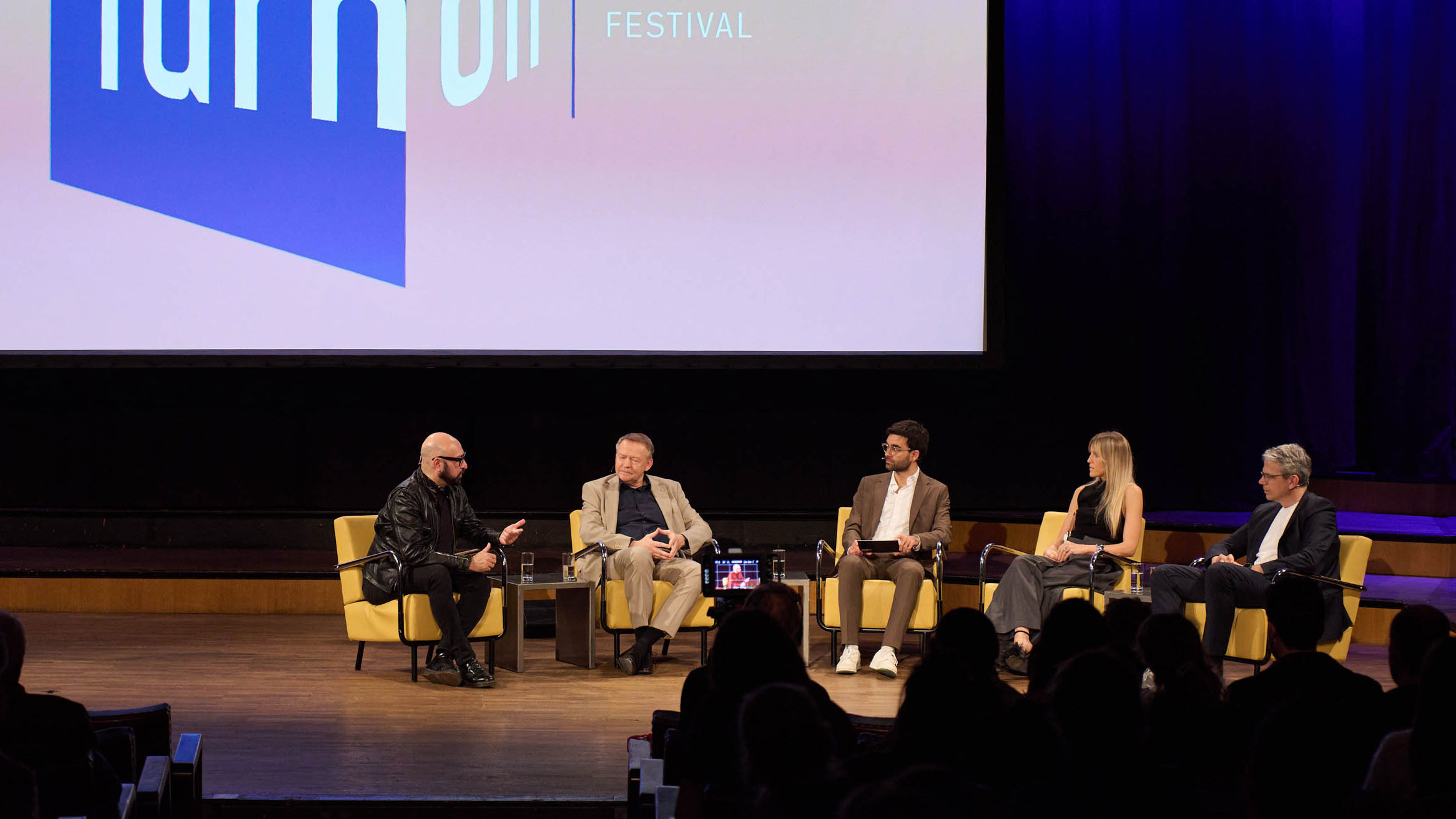 Stage panel of five speakers in yellow chairs; large screen displays festival branding.
