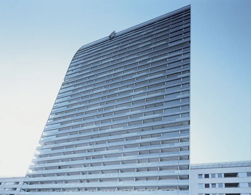 Tall glass high-rise residential building with multiple balconies, viewed from below against a blue sky.