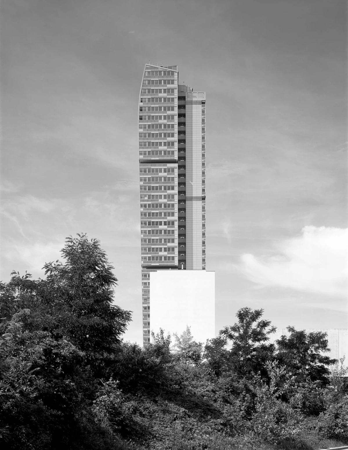 Tall modern high-rise tower with a curved top rising from a white base, surrounded by trees.