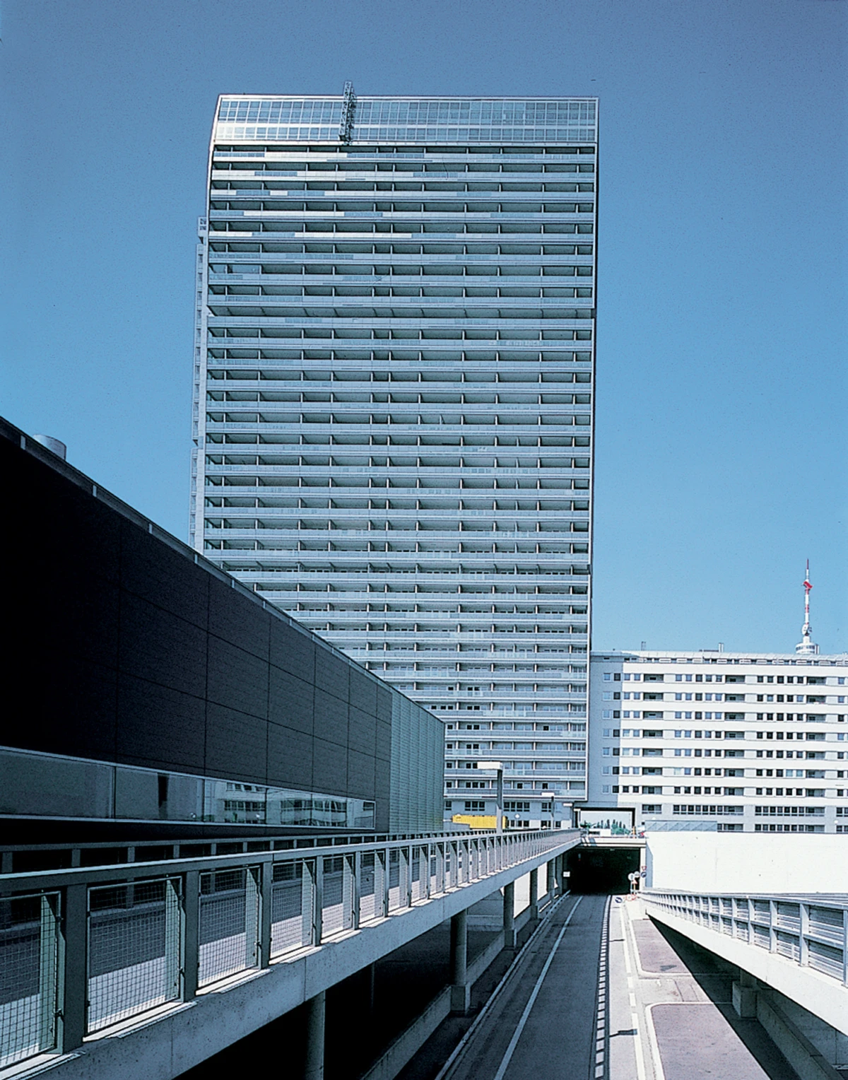 Glass-clad high-rise towers over an urban skyline; an elevated roadway with railings leads to an underground tunnel.