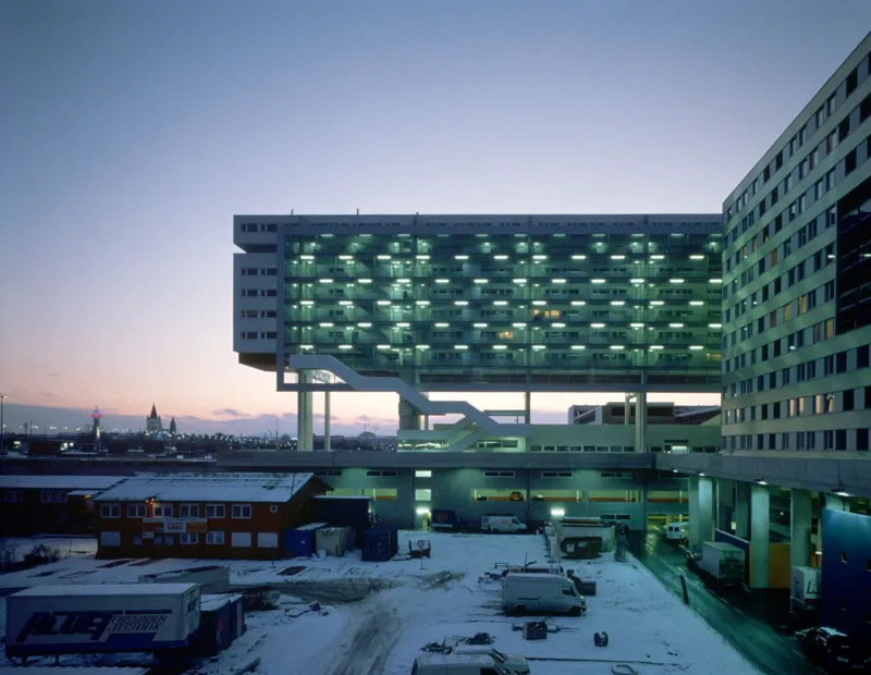 Modern hospital complex with a glass-walled elevated block atop a platform; dusk, snow-covered loading yard and trucks.