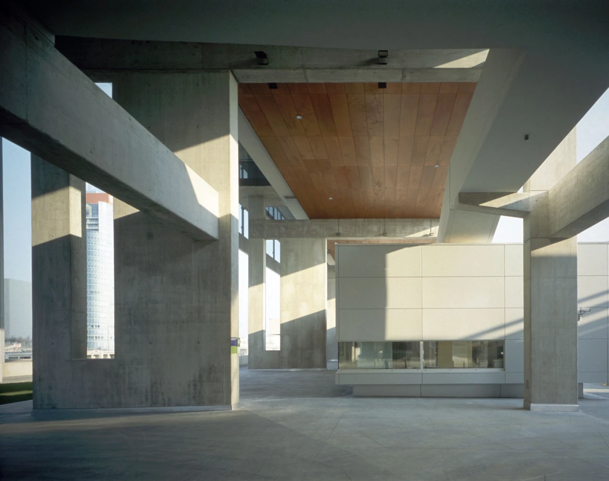 Empty outdoor space under a concrete elevated structure; large columns, wooden ceiling, and a modern rectangular building.