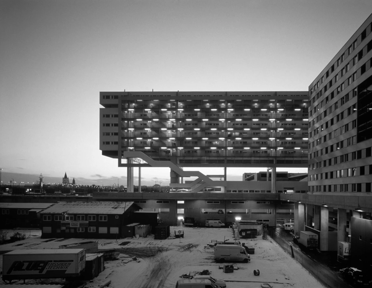 Elevated modern parking garage on stilts connected to a multi-story office building, snow-covered lot with vehicles at dusk.