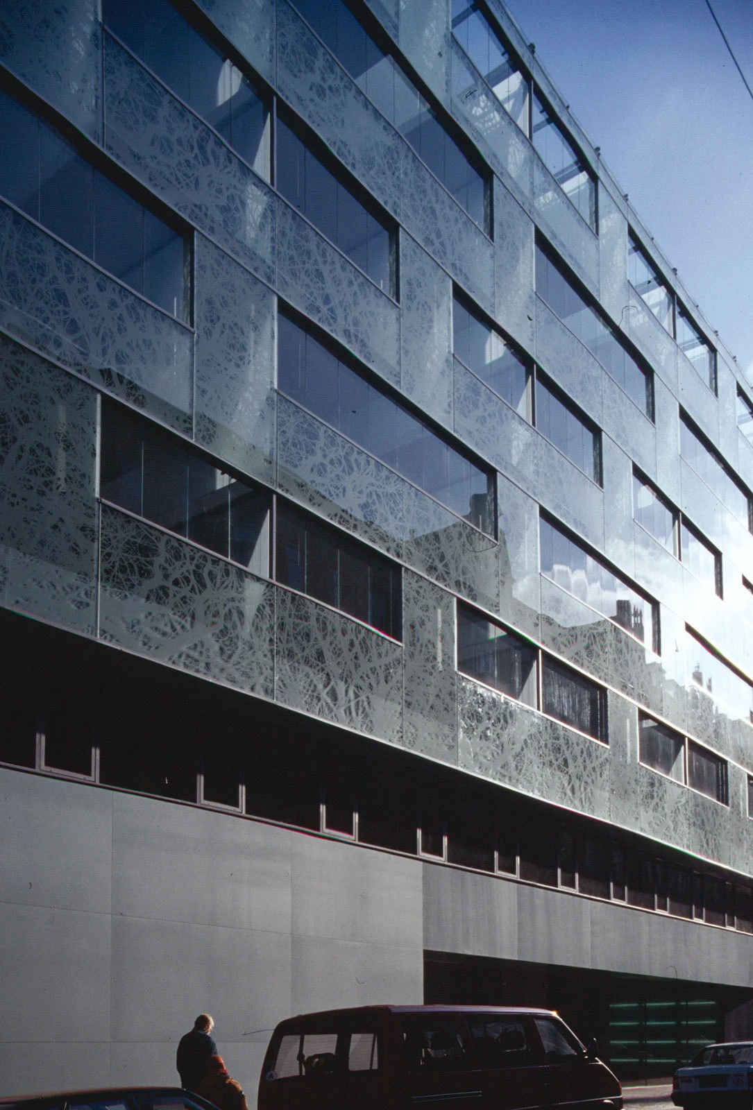 Glass-clad multi-story office building with a patterned facade; a person walks beside a dark red van at street level.