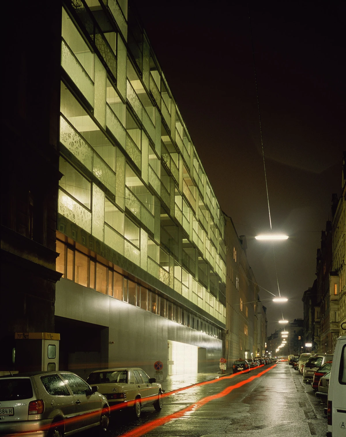 Glass-fronted building at night along a wet city street; red light trails from passing cars.