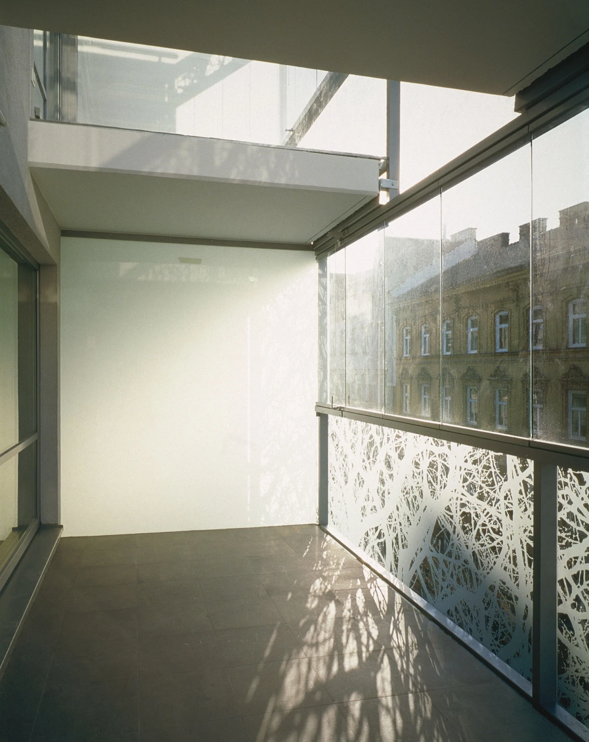 Glass-walled balcony with tiled floor; sunlit shadows on the floor, white wall at the far end, and a brick building visible through the glass railing.