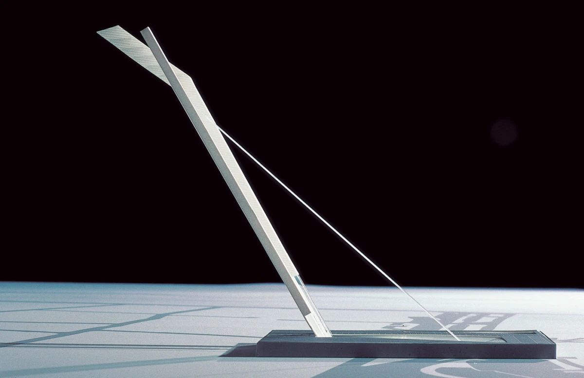 Metal inclinometer on a rectangular base, angled with a taut wire, set on a pale surface against a black background.