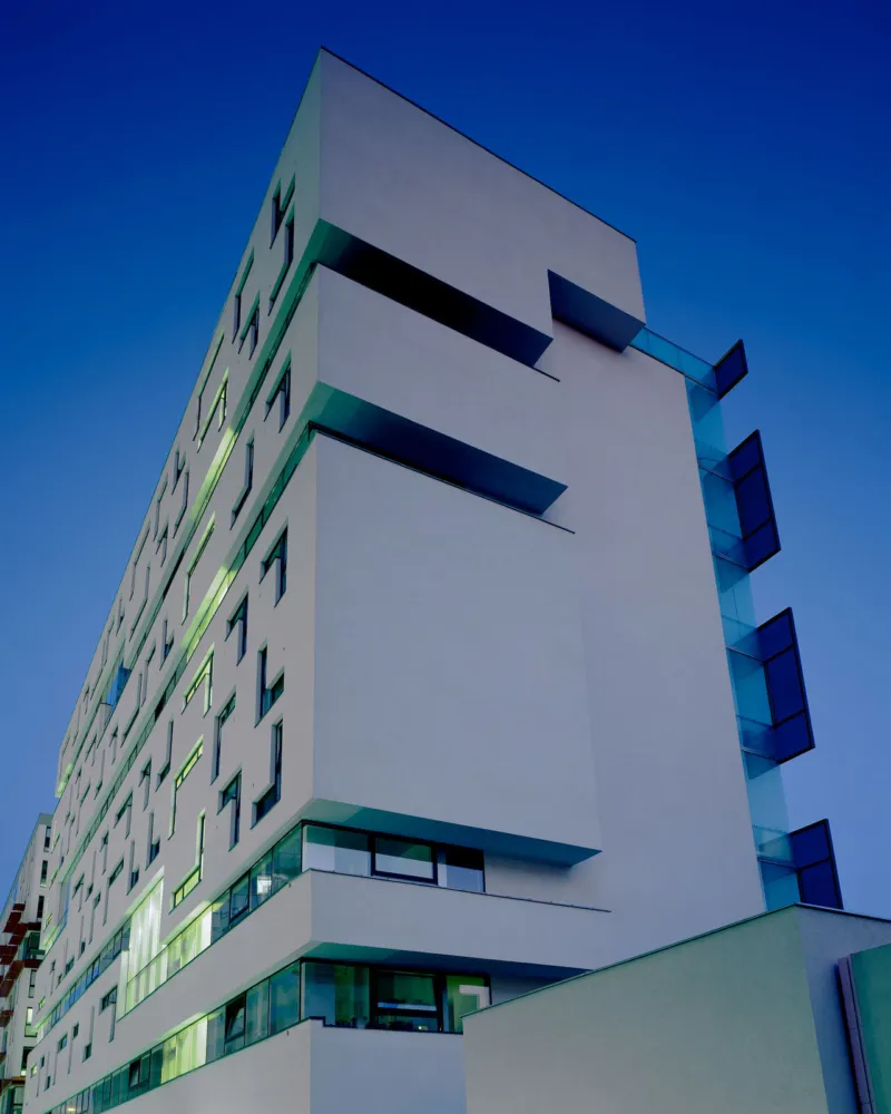 Urban modern white apartment building, photographed from ground looking up, with glass balconies against a clear blue sky.
