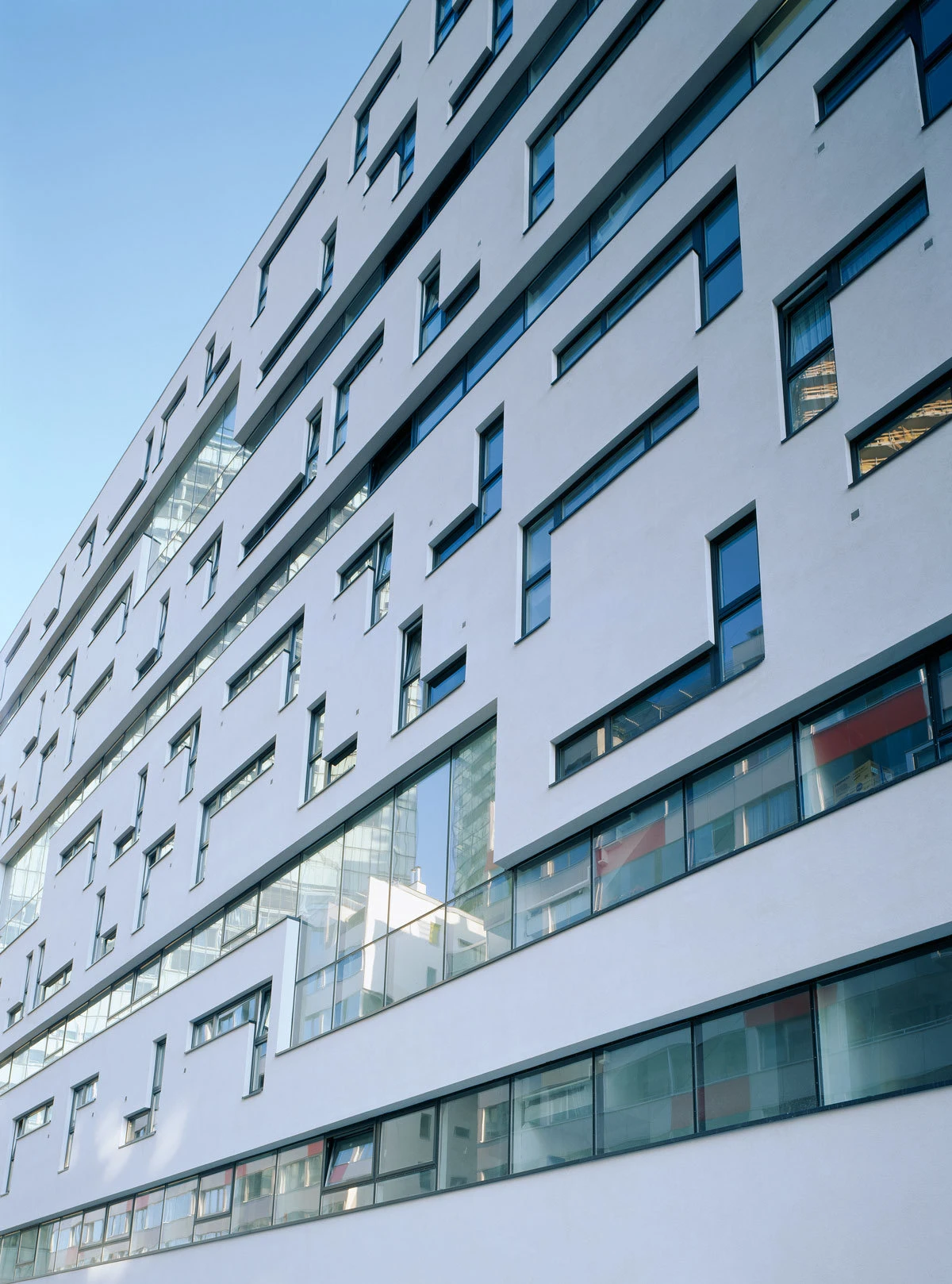 Diagonal-perspective view of a white modern building facade with irregular, staggered rectangular windows against a blue sky.