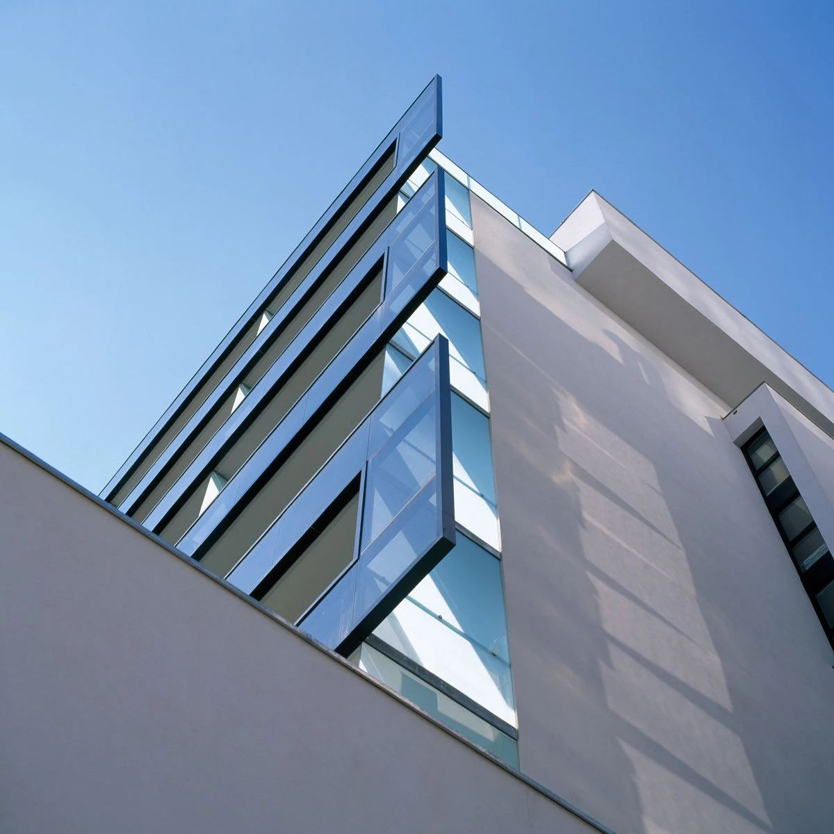 Corner of a contemporary glass-clad building with cantilevered glass balconies, set against a clear blue sky.