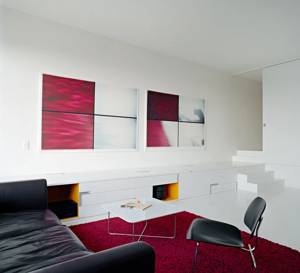 Minimalist living room with white walls and cabinetry; pink abstract diptych above, black leather sofa, white coffee table, black chair, red rug.