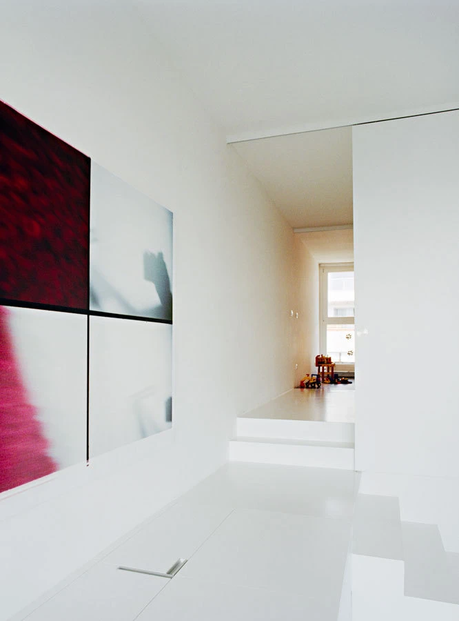 Bright white modern hallway with four-panel abstract artwork on the left; glossy white stairs lead to a sunlit room with toys at the far end.