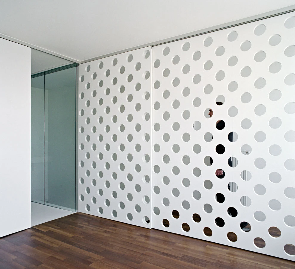 White perforated wall panel with circular holes; silhouettes visible through openings in a modern interior with glass door and wooden floor.