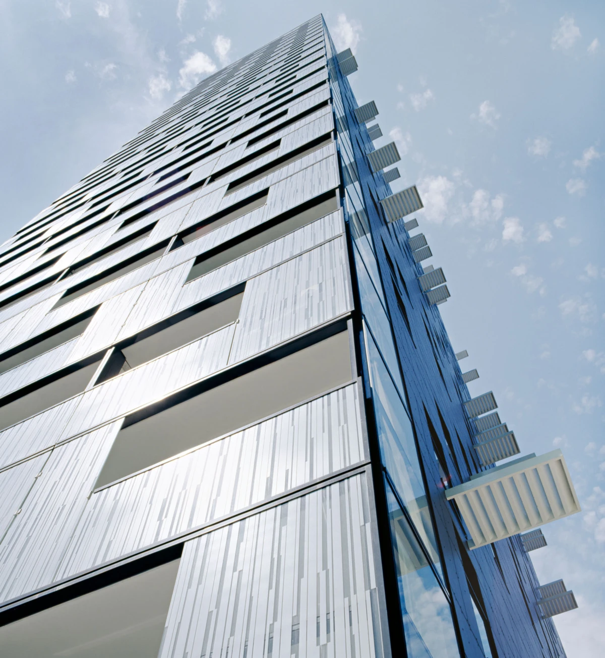 Low-angle view of a modern glass high-rise with vertical panels and white cantilevered sunshades against a blue sky.