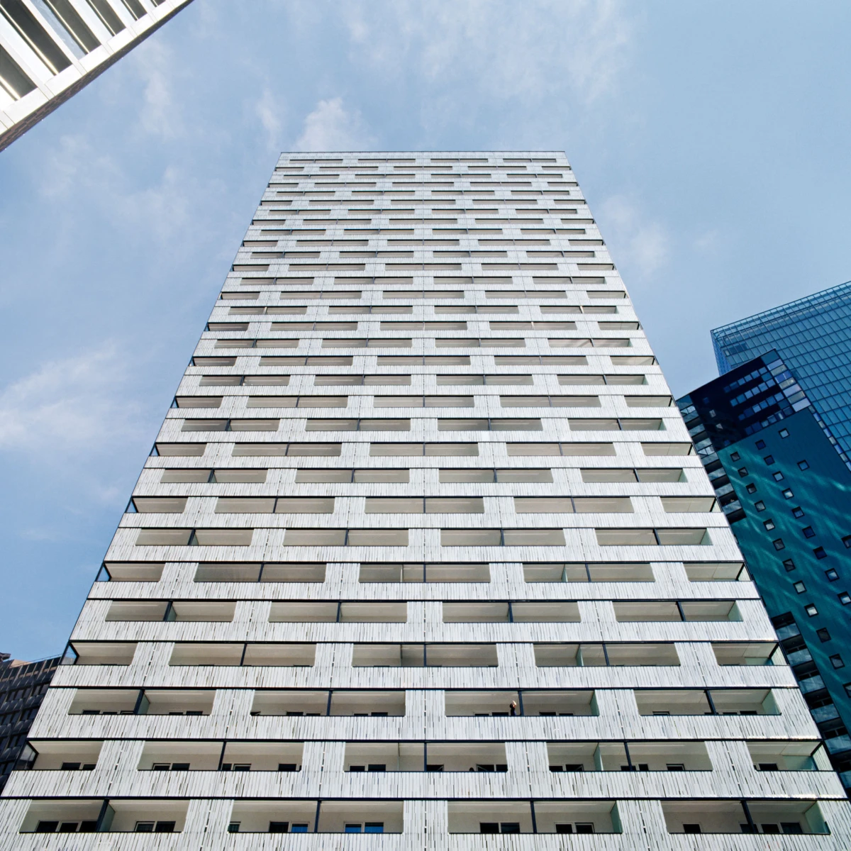 Tall white apartment building rising with repetitive balconies, viewed from street level against a blue sky.