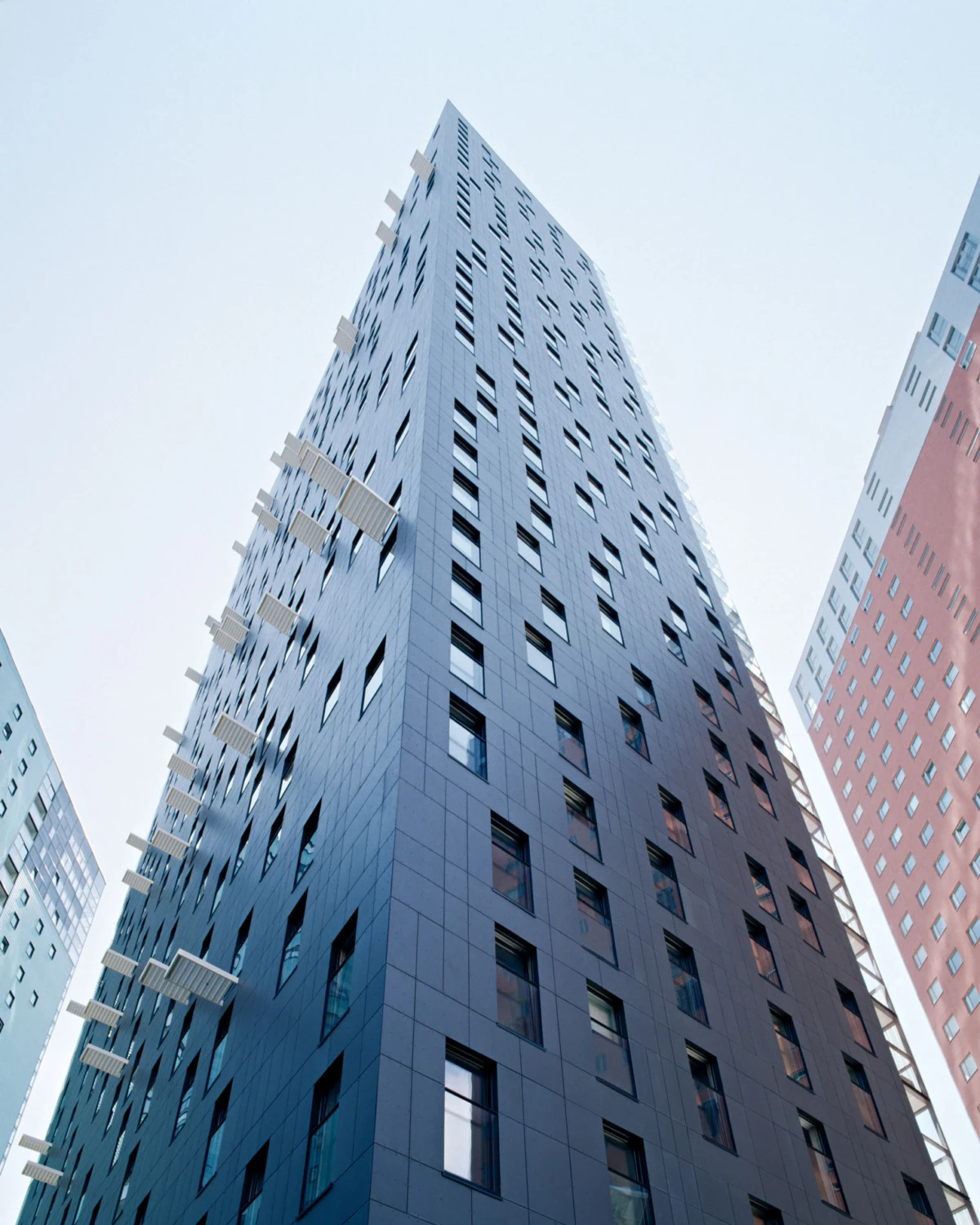 Blue-gray high-rise building seen from street in an urban cityscape, with numerous square windows and protruding balconies.
