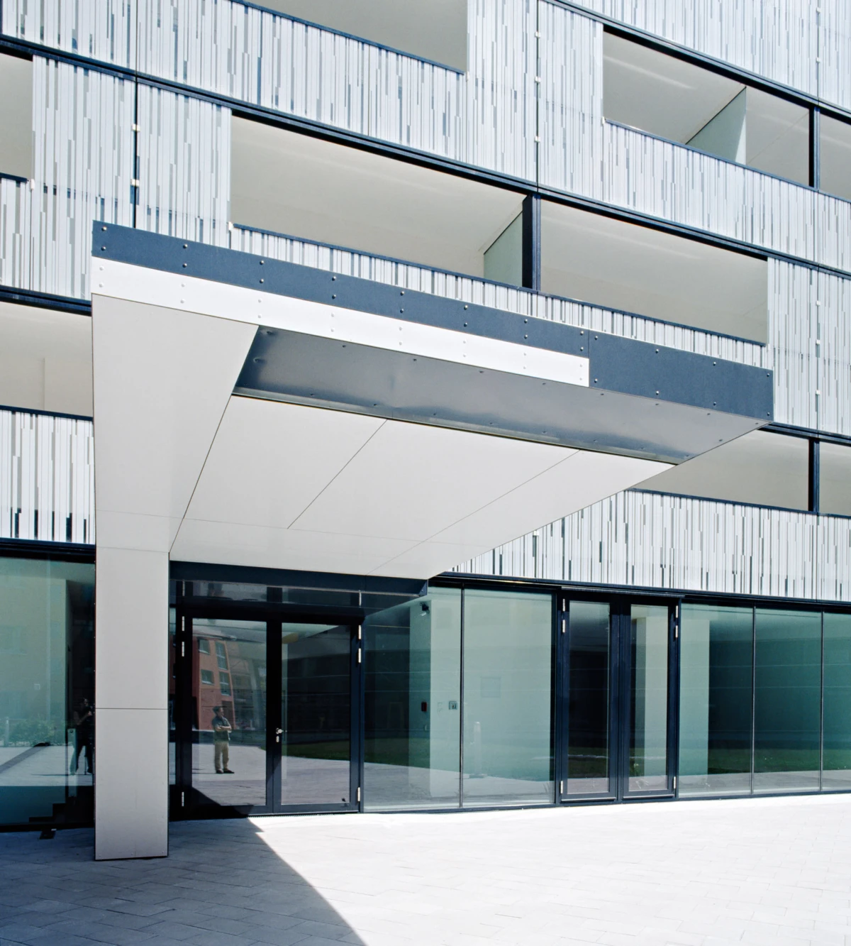 Modern office building entrance with glass doors under a white cantilevered canopy; metal and glass facade and paved plaza.