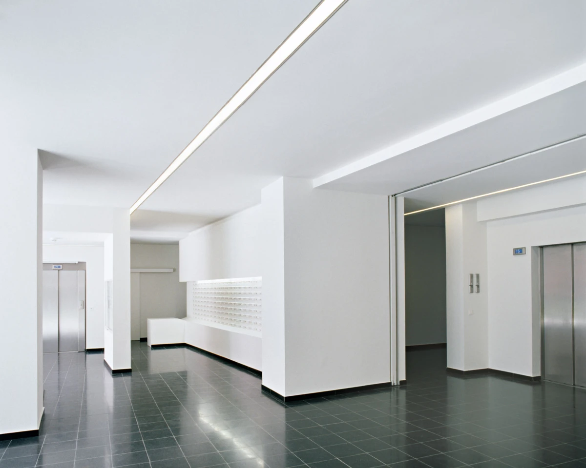 Modern empty lobby with white walls, dark tile floor, and elevator doors at opposite ends; linear ceiling lights and a white bench along the wall.