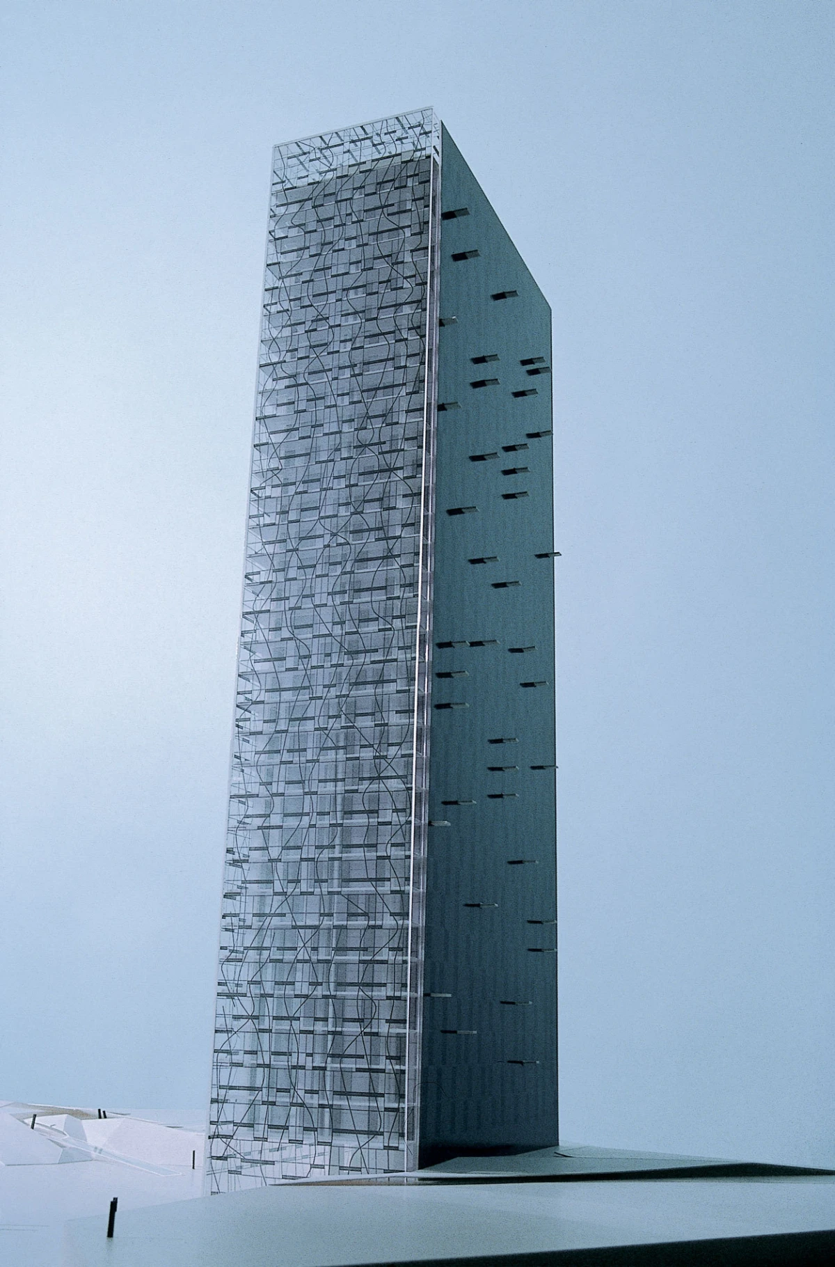 Tall slender skyscraper with lattice-pattern glass facade beside a solid blue wall under a pale blue sky.