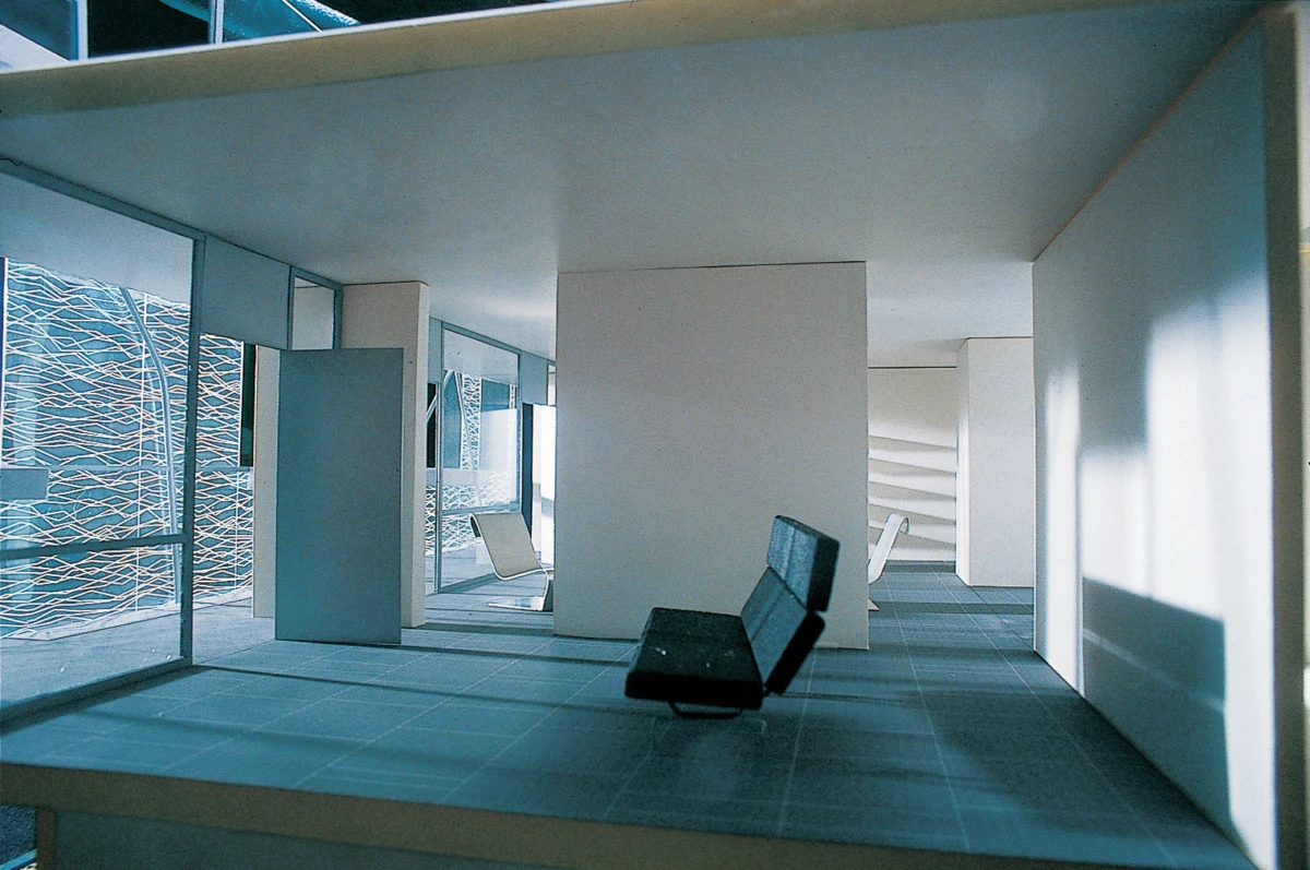 Scale model of a modern interior with white partition walls, a black sofa, and a glass facade on the left; blue daylight fills the space.