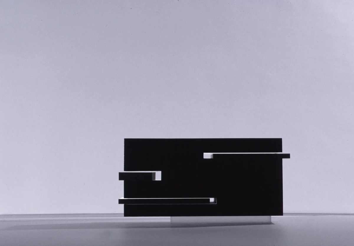 Abstract black rectangular sculpture with stacked blocks and white protruding slats, set against a pale studio backdrop.