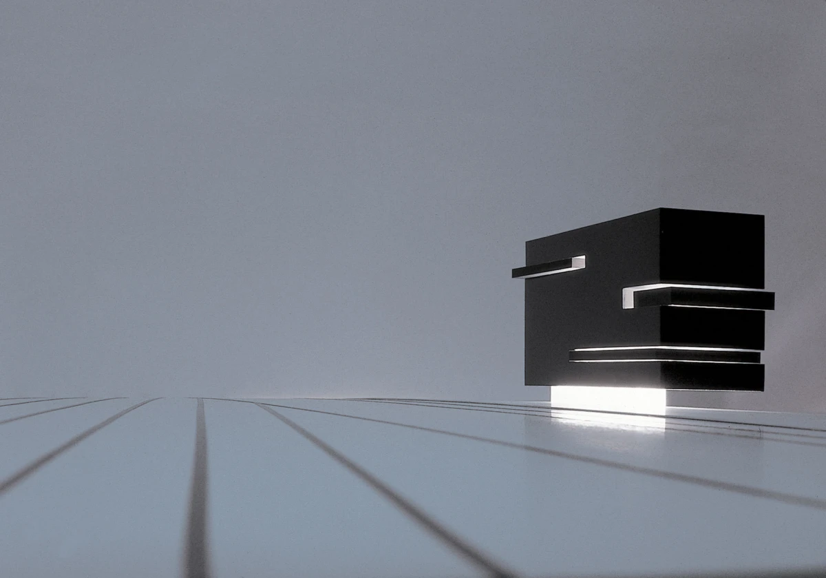 Black geometric sculpture of stacked rectangular blocks on a glossy, reflective surface with diagonal lines and a gray backdrop.