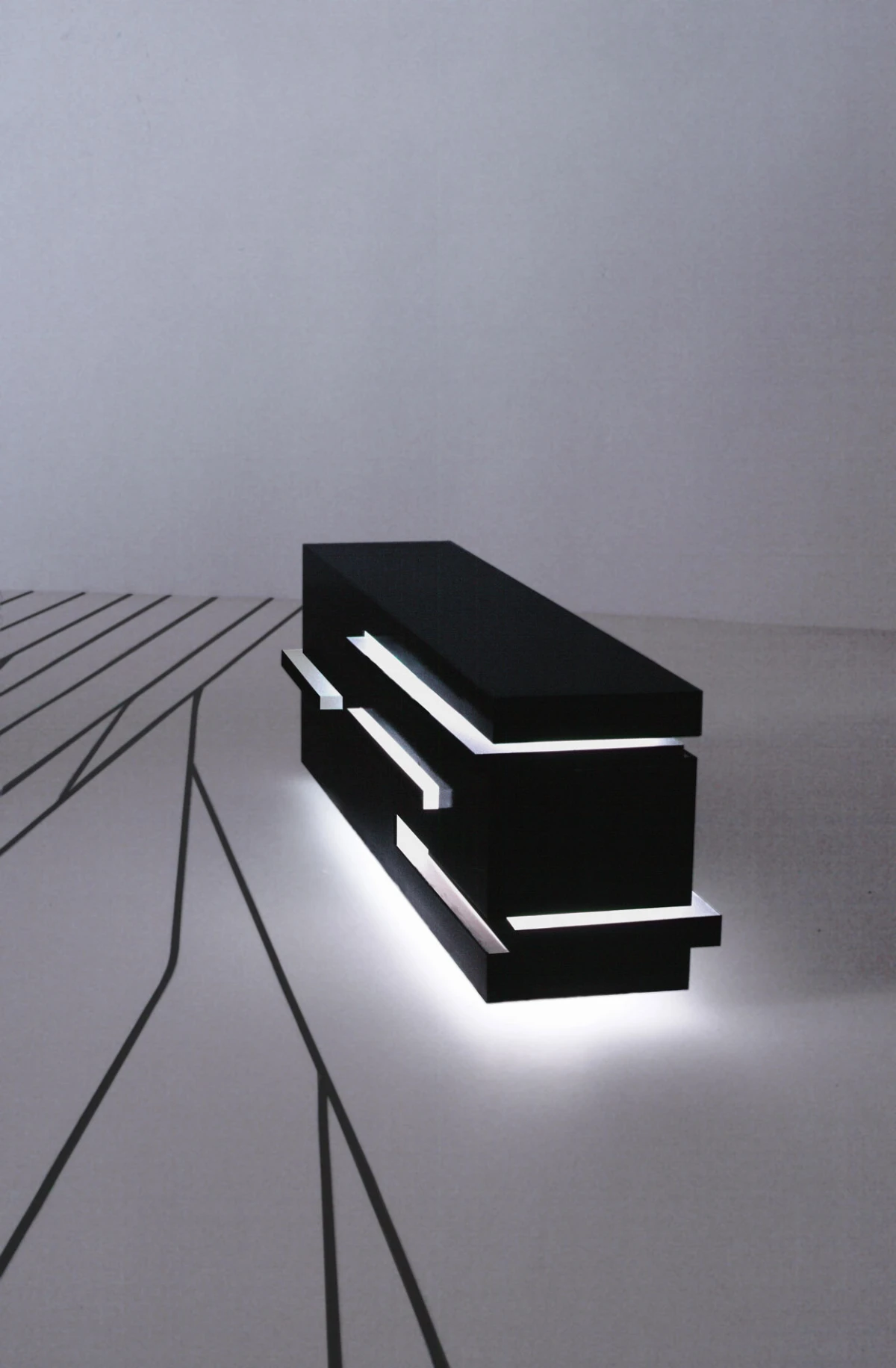 Black two-tier rectangular block sculpture with glowing white edges on a white reflective surface.