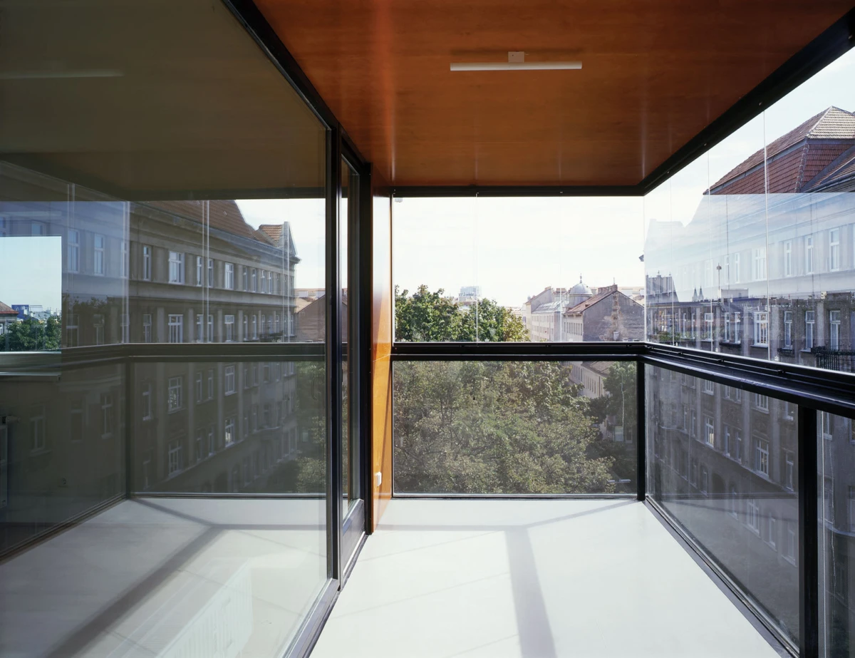 Unoccupied glass-walled balcony with metal railing and wooden ceiling overlooking a tree-lined city street.