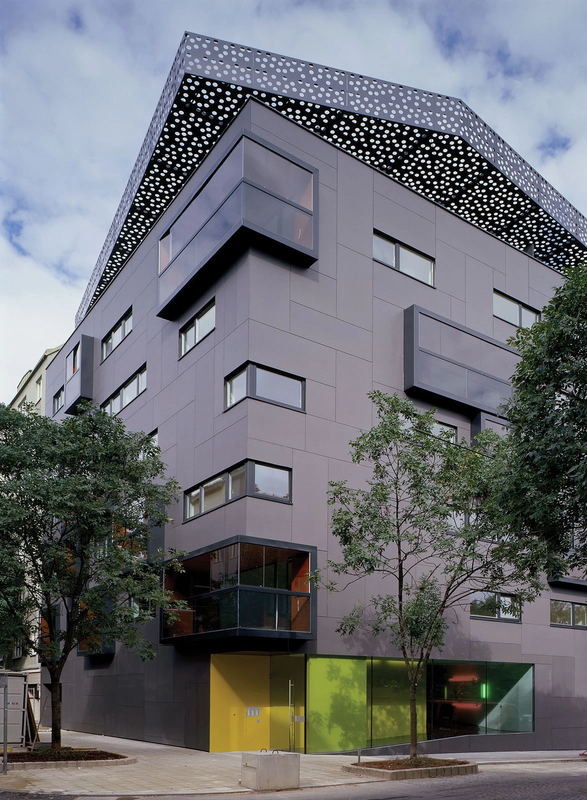 Modern gray apartment building with cantilevered balconies, perforated roof canopy, and yellow entry, beside trees on a city street.