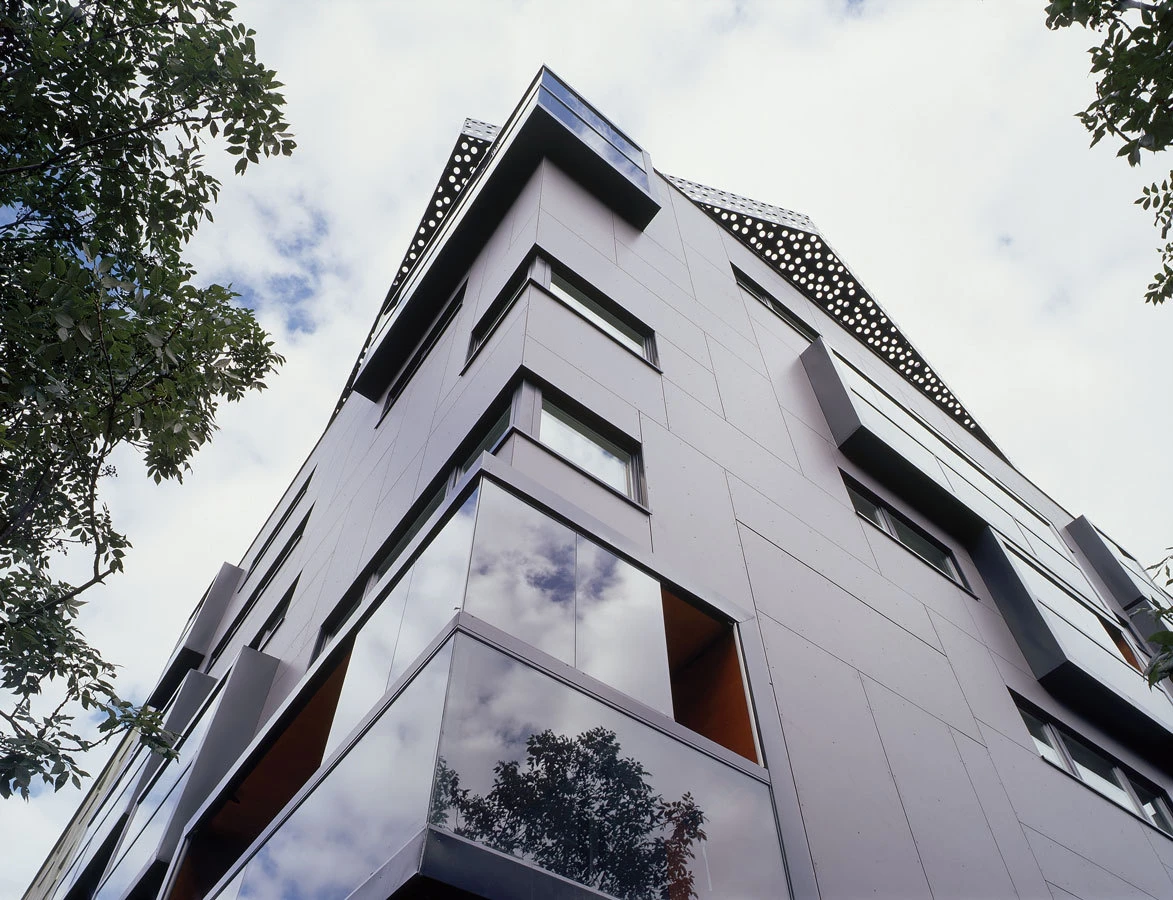 Low-angle view of a modern gray apartment building with glass balconies and sharp windows, framed by trees under a cloudy sky.