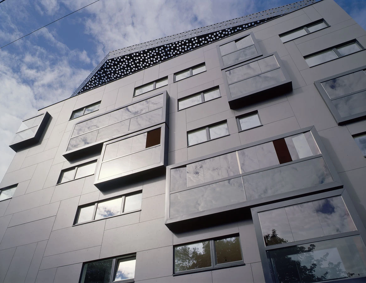 Modern gray apartment building with staggered rectangular balconies, metal panels, and reflective windows under a cloudy sky.
