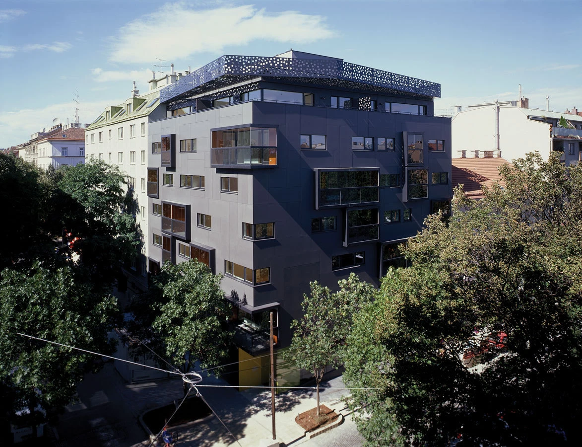Dark gray multi-story residential building with protruding balconies on a tree-lined city street; perforated lattice rooftop.