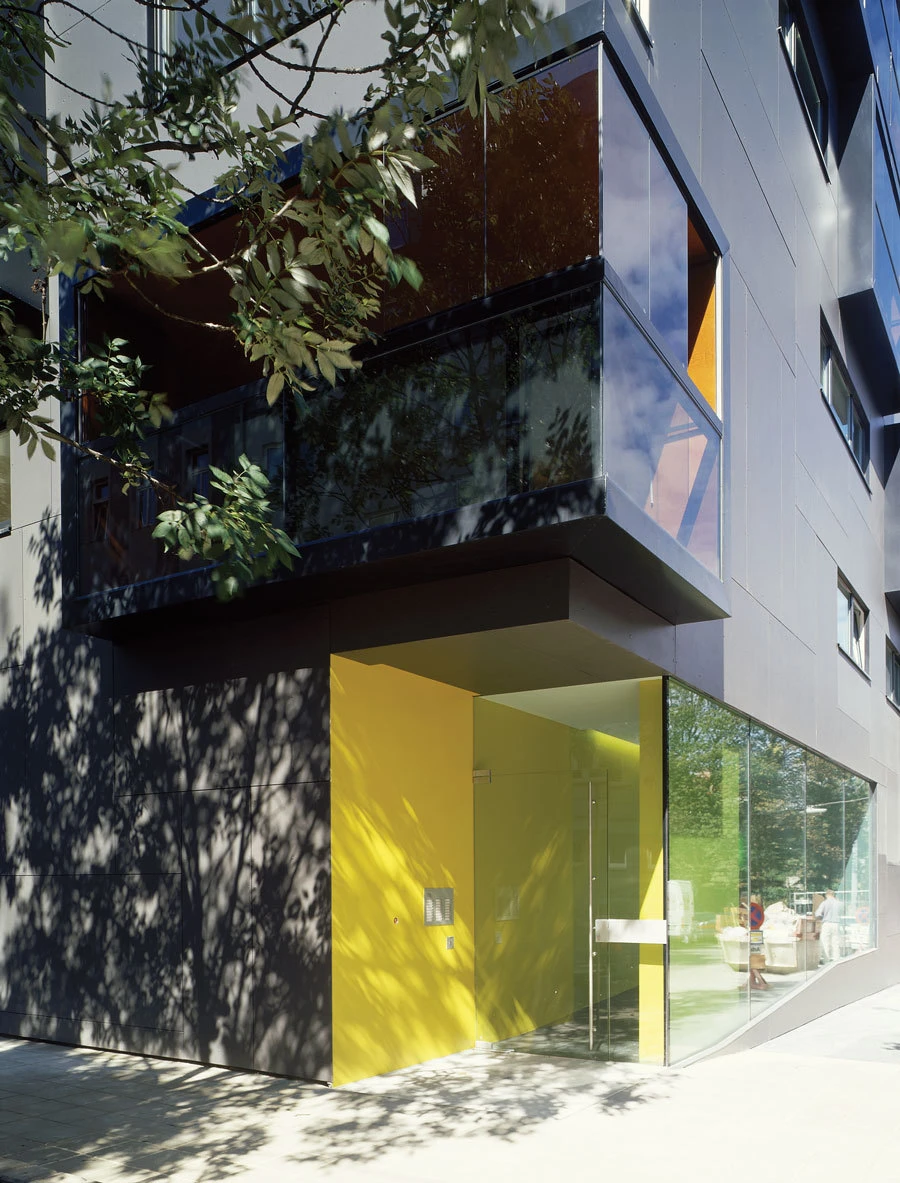 Modern building entrance with glass doors and a bright yellow wall panel; dark facade and tree-shadowed sidewalk.