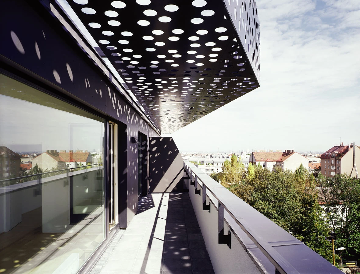 Balcony with perforated metal canopy overhead casting circular shadows, overlooking a city skyline.