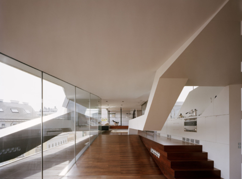 Contemporary open-plan interior with glass wall along the left, dark wooden staircase on the right, and a white kitchen at the far end.