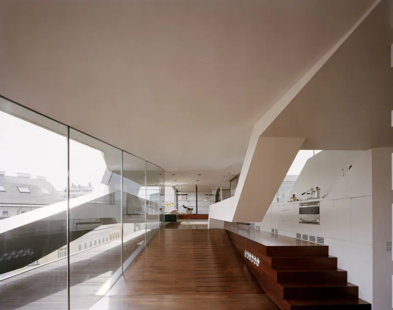 Contemporary open-plan interior with glass wall along the left, dark wooden staircase on the right, and a white kitchen at the far end.