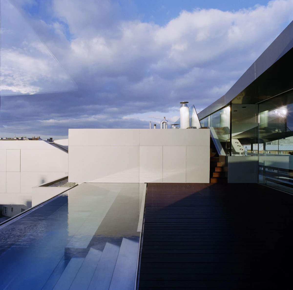 Modern rooftop terrace with white concrete walls, glass-walled extension, wooden deck, and a reflective pool along the edge.