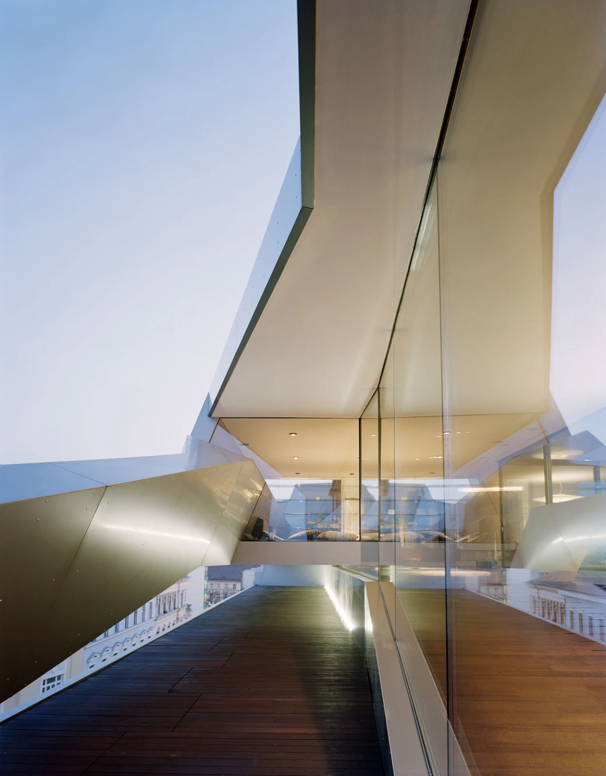 Contemporary glass-walled building with angular metallic overhangs, a wooden deck walkway, and a distant cityscape.