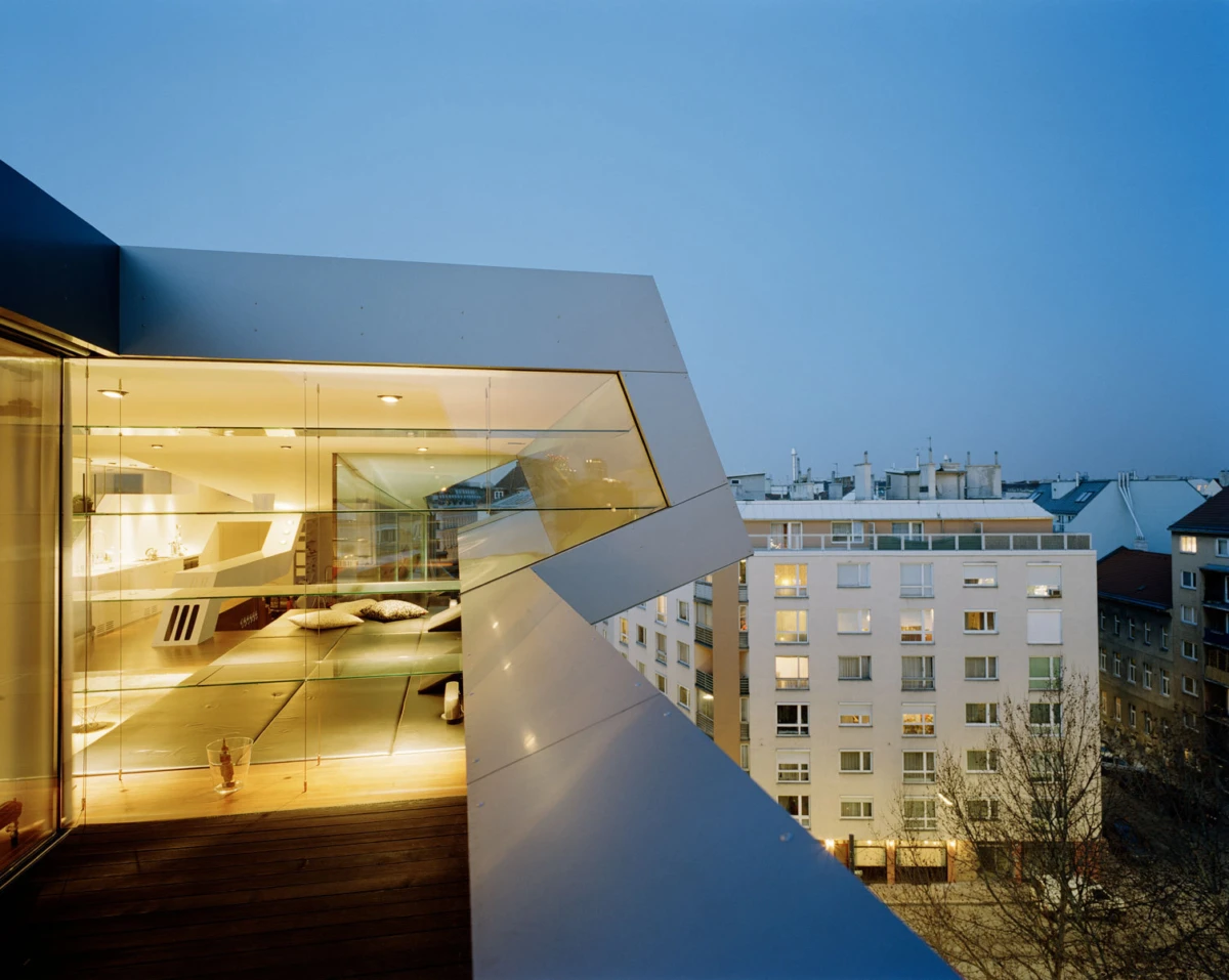Glass-walled rooftop living area with wooden deck, illuminated interior, overlooking city apartment buildings at dusk.