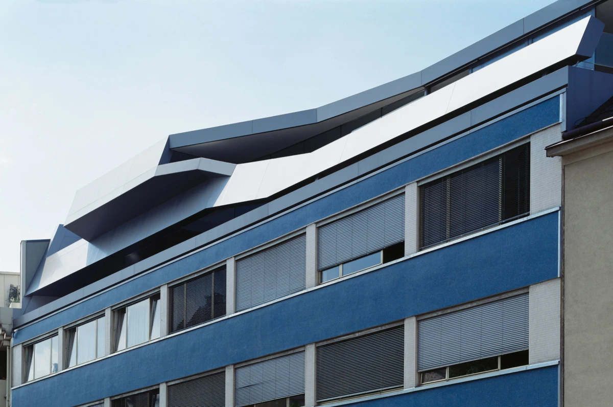 Blue multi-story office building with angular white rooftop structures and windows with blinds.