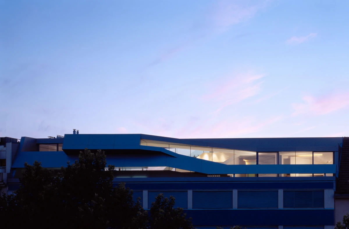 Blue modern office building with a curved top-floor band and glass facade at dusk; trees in foreground.