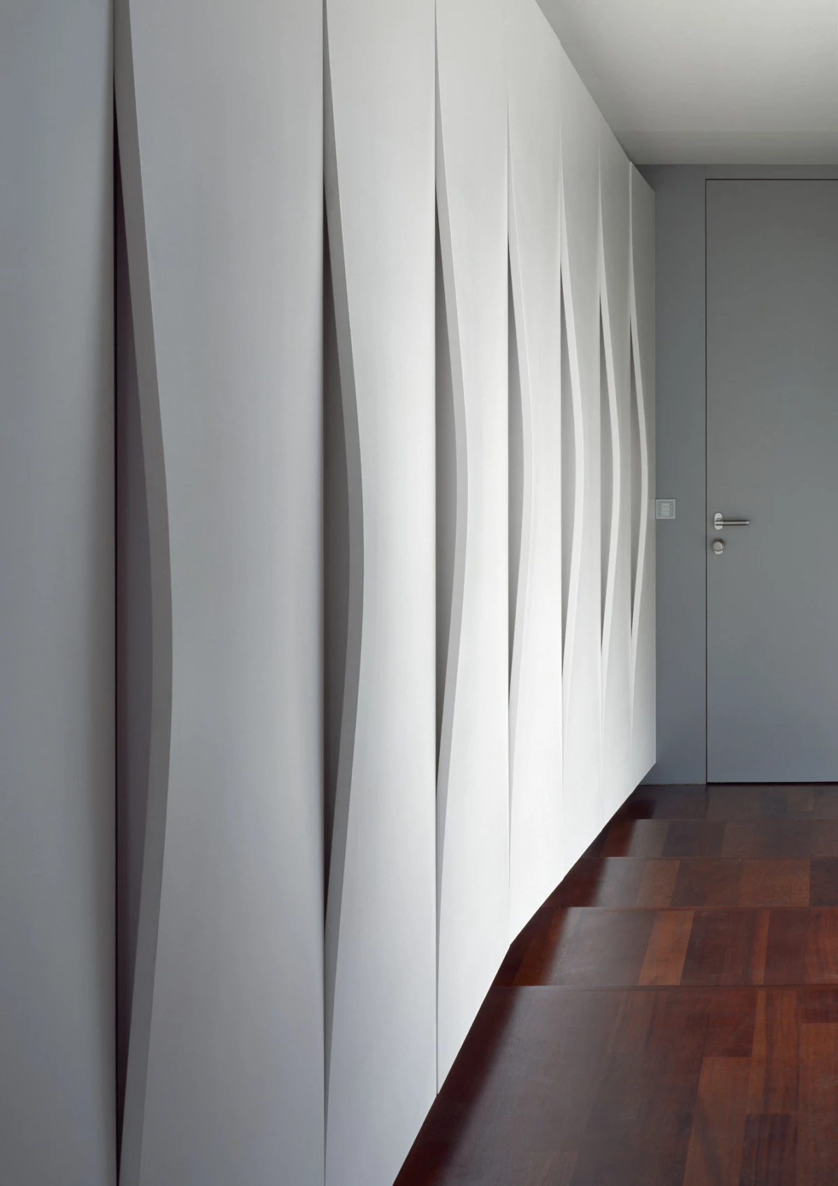 White undulating 3D wall panels line a hallway with a gray door and dark hardwood floor.