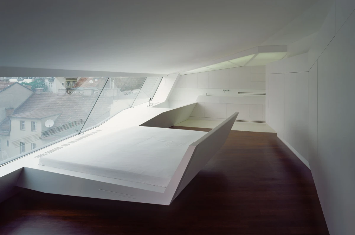 Minimalist white loft with angular platform bed, built-in cabinetry, skylights, and dark wood floor; city rooftops visible through large windows.