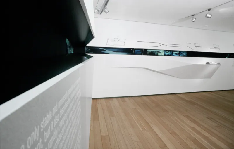 Contemporary art gallery interior with a white angular display wall, a horizontal strip of images, and a wooden floor.