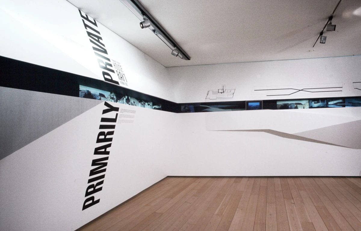 Interior of a contemporary art gallery with white angular walls, a black horizontal band, light wood floor, and bold vertical text along the wall.